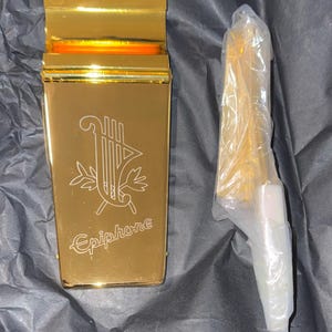 May include: A gold-colored metal guitar pick holder with the Epiphone logo and a lyre design. The holder is packaged in a clear plastic bag.