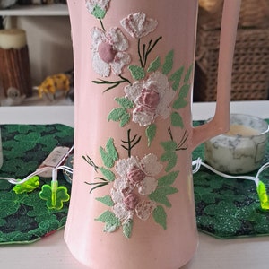 May include: A pink ceramic pitcher with a floral design. The flowers are white with pink centers and green leaves. The pitcher has a lid and a handle.