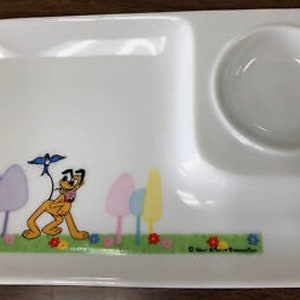 May include: A white divided plate with a cartoon image of Pluto the dog, a blue bird, and colorful trees. The plate has a small, round, white bowl attached to the side.