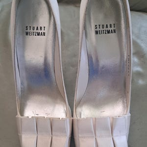 May include: Pair of off-white Stuart Weitzman high heels. The open-toe shoes have a pleated fabric detail across the top and a silver insole. The brand name is printed in black on the insole. The heels are approximately 3 inches high.