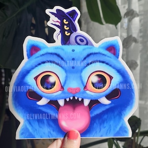 May include: A blue cat sticker with large orange eyes, white fangs, and a pink tongue. The cat has a hat and a small design on its head. The sticker is held by a hand. The website oliviaoltmanns.com is printed on the sticker.