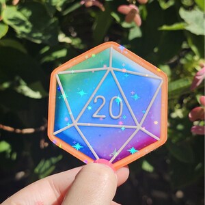 May include: A colorful sticker of a twenty-sided die, with a rainbow gradient and the number 20 in the center. The die has an orange border and is decorated with small star-shaped sparkles.