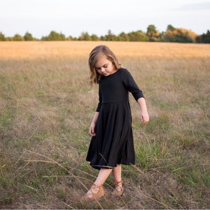 May include: A young girl in a black dress walks through a field of tall grass. She is looking down at the ground and her hair is long and curly.