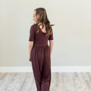 May include: A burgundy jumpsuit with short sleeves and a gathered waist. The model is facing away from the camera, standing on a light-coloured wooden floor. The background is a plain, light-coloured wall.
