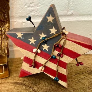 May include: A five-pointed wooden star ornament painted with a red, white, and blue American flag pattern. The star is decorated with thin, brown, wire-wrapped stems featuring small, round, red, white, and blue berries attached to the front.