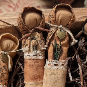 May include: Close-up of several handmade fabric dolls. The dolls have tan bodies and patterned head coverings. Each doll has a unique embellishment, including lace, a small heart charm, and dried flowers. The dolls are arranged in a rustic setting.