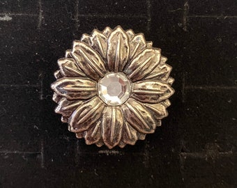 May include: A silver-toned, flower-shaped button with a clear, faceted gem in the center. The button has detailed petal-like edges and is set against a dark background.