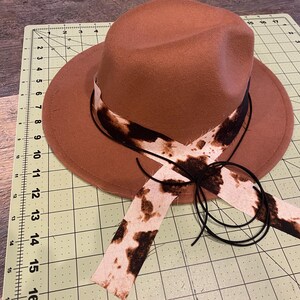 May include: A brown fedora hat with a black and white cow print ribbon. The hat has a black string tied around it.