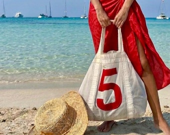 May include: A white canvas tote bag featuring a large red number 5 stitched on the front, paired with a natural tan straw hat. The bag has two long shoulder straps and visible white stitching details along the seams of the fabric construction.