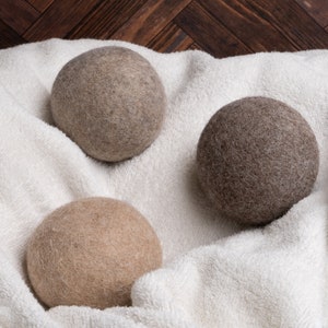 May include: Three wool dryer balls in different shades of brown, beige, and gray, resting on a white towel.