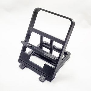 May include: A black plastic phone stand with a raised platform for holding a phone. The stand has a simple design and is angled for viewing.
