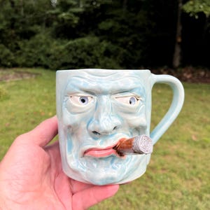 May include: A light blue ceramic mug featuring a sculpted, three-dimensional human face with a stern expression. The face has detailed eyes, a nose, and pink lips, with a brown cigar protruding from the mouth. The mug includes a curved handle.