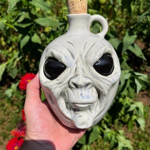 May include: A white ceramic alien head jug with a cork stopper. The jug has black eyes and a wide mouth.
