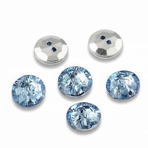 May include: A selection of decorative buttons. The image shows five light blue, faceted crystal buttons and two silver-coloured buttons with two holes each. The buttons are arranged on a white background.