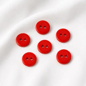 Könnte beinhalten: Sechs kleine, runde, leuchtend rote Knöpfe, die jeweils zwei mittige Löcher zum Annähen aufweisen. Diese kreisförmigen Verschlüsse haben eine glatte, einfarbige Oberfläche und sind verstreut auf einem weißen Stoffhintergrund angeordnet.