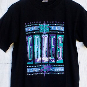 Peut inclure: T-shirt noir avec un imprimé graphique du logo des Jeux d'hiver de 1995 en Colombie-Britannique. Le logo comprend le texte "British Columbia Winter 95 Games" et une chaîne de montagnes stylisée avec un éclair.