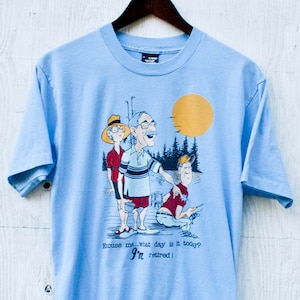 May include: Light blue t-shirt featuring a cartoon illustration of three people fishing. The text on the shirt reads "Excuse me, what day is it today? I'm retired!"