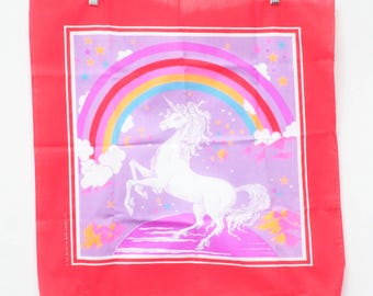May include: A red square textile with a white unicorn graphic, rainbow, and stars. The unicorn is standing on a pink surface, with a purple background. The textile is hanging on a wooden hanger.