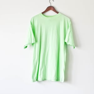 Op de afbeelding: Een lichtgroen T-shirt hangt aan een houten hanger tegen een witte achtergrond.
