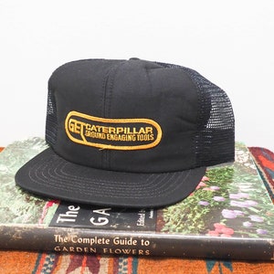 Könnte beinhalten: Schwarze und gelbe Trucker-Kappe mit einem Caterpillar-Logo, auf dem "Get Caterpillar Ground Engaging Tools" steht. Die Kappe liegt auf einem Buch und einem gewebten Teppich.