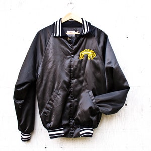 Puede incluir: Una chaqueta de béisbol de satén negro con ribetes de rayas blancas y negras en el cuello, los puños y el dobladillo. La chaqueta tiene un diseño bordado amarillo en el pecho izquierdo.