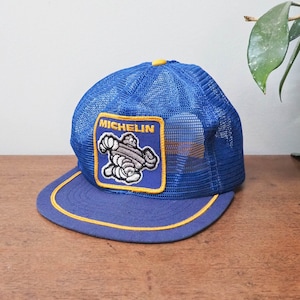 May include: Blue mesh trucker hat with a yellow brim and a blue and yellow Michelin Man patch. The patch has the word "MICHELIN" in white letters.