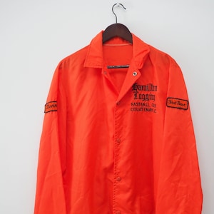 Puede incluir: Chaqueta de nylon naranja con cremallera negra y botones a presión. La chaqueta tiene bordado "Hamilton Logging Fastball Club Courtenay, B.C." en la parte delantera y parches "First Base" y "Denn" en las mangas.