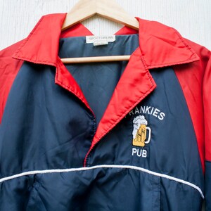May include: A red and navy blue windbreaker jacket with a white stripe. The jacket has an embroidered logo that says "Frankies Pub" with a beer mug graphic.