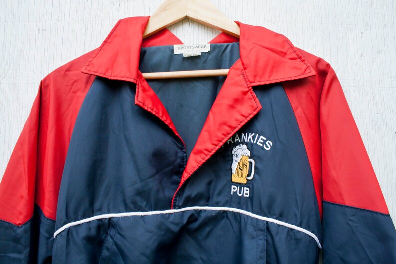 May include: A red and navy blue windbreaker jacket with a white stripe. The jacket has an embroidered logo that says "Frankies Pub" with a beer mug graphic.