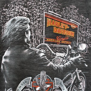 May include: A dark grey t-shirt with a graphic print of a person riding a motorcycle. The graphic includes a sign that reads "Harley-Davidson Genuine American Motorcy" and a banner that reads "Since 1903 Follow Nobody".