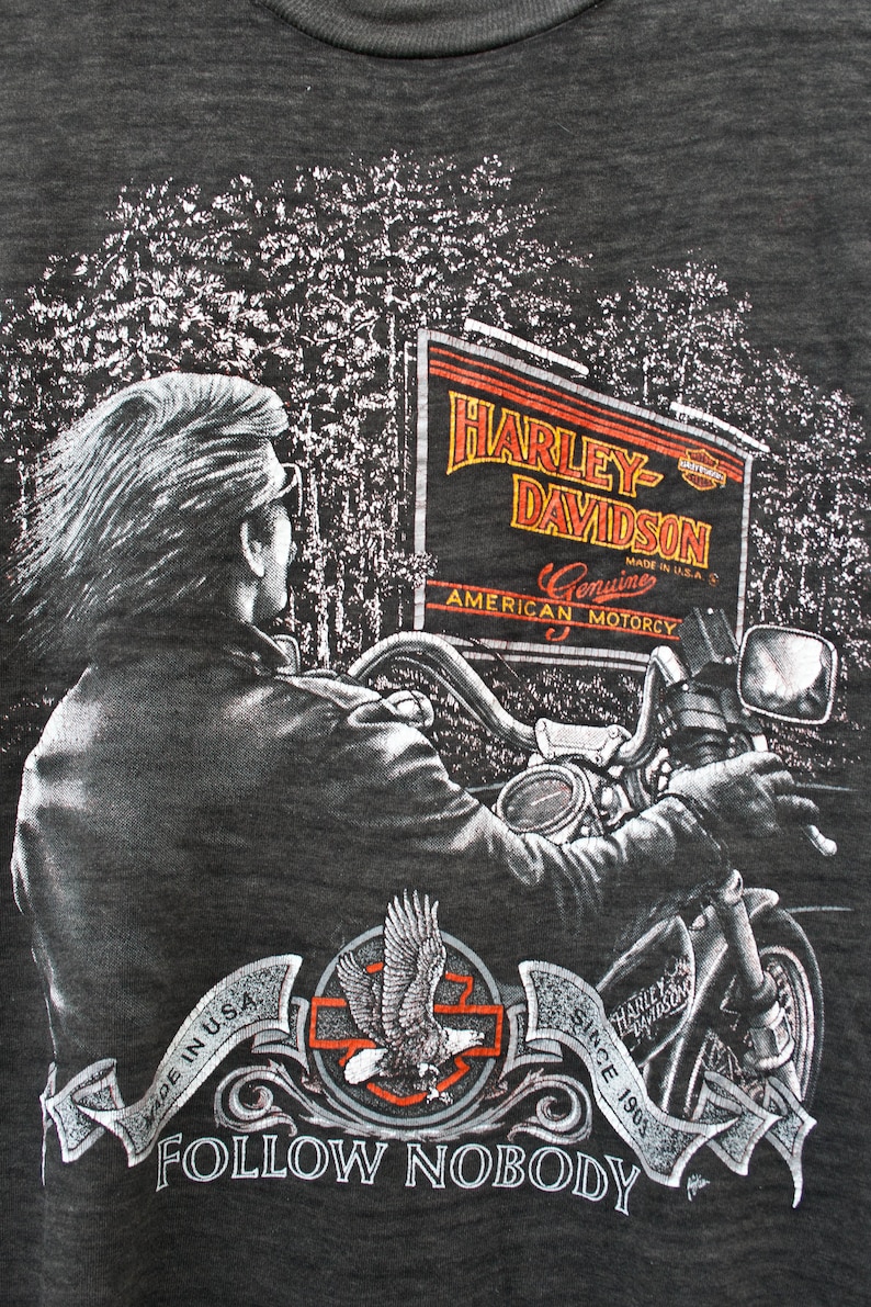 May include: A dark grey t-shirt with a graphic print of a person riding a motorcycle. The graphic includes a sign that reads "Harley-Davidson Genuine American Motorcy" and a banner that reads "Since 1903 Follow Nobody".