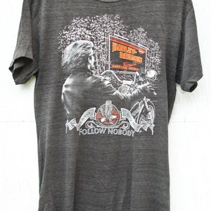 May include: A gray t-shirt with a Harley-Davidson graphic. The graphic features a motorcycle rider in a forest setting with a sign that reads "Harley-Davidson, Genuine American Motorcycle". The bottom of the graphic reads "In U.S.A. Since 1903, Follow Nobody".