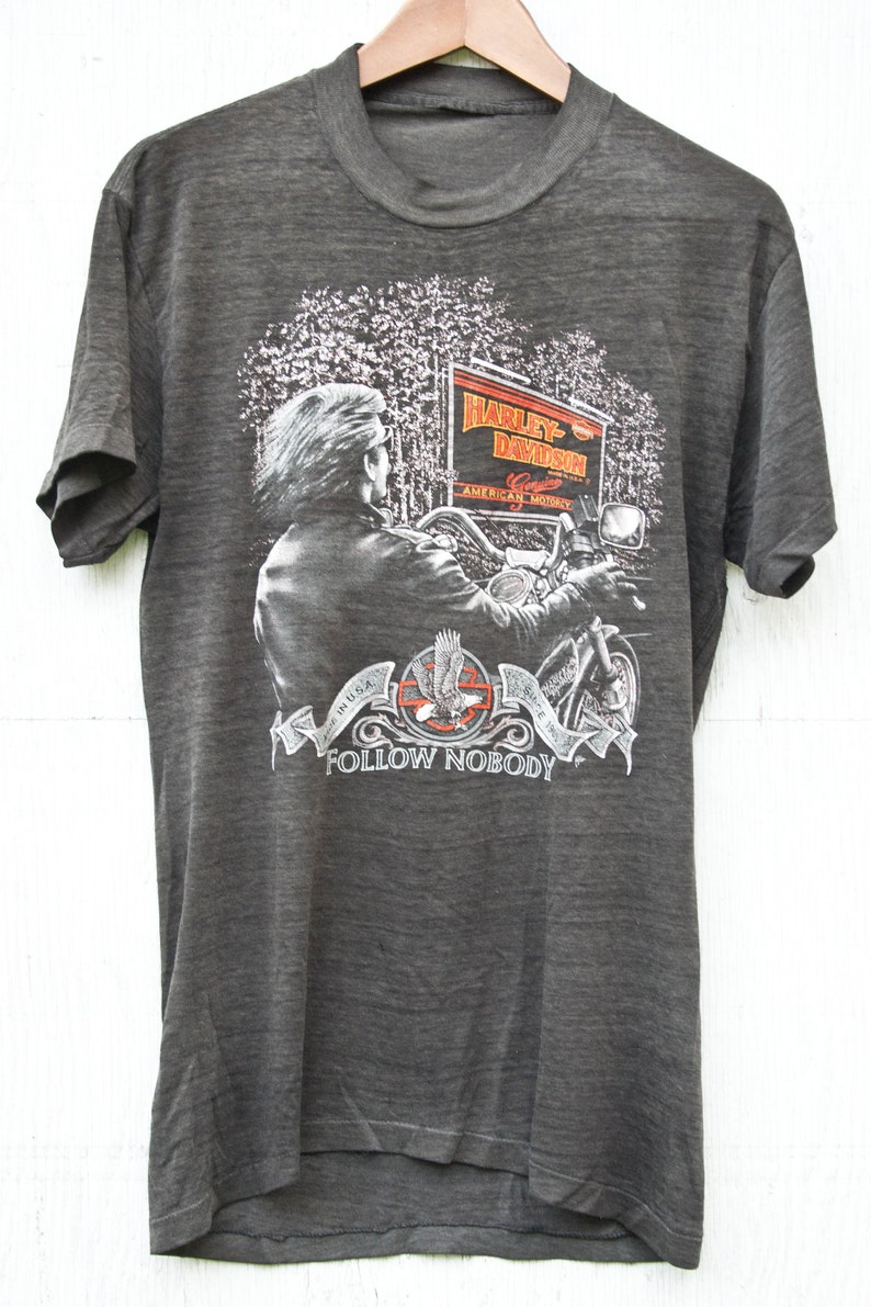 May include: A gray t-shirt with a Harley-Davidson graphic. The graphic features a motorcycle rider in a forest setting with a sign that reads "Harley-Davidson, Genuine American Motorcycle". The bottom of the graphic reads "In U.S.A. Since 1903, Follow Nobody".