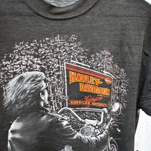 May include: A gray t-shirt with a Harley-Davidson graphic featuring a motorcycle rider in a forest setting. The graphic includes the text "Harley-Davidson", "Genuine American Motorcy", and "Since 1903". The shirt also has the text "Made in USA" and "Follow Nobody" printed on it.