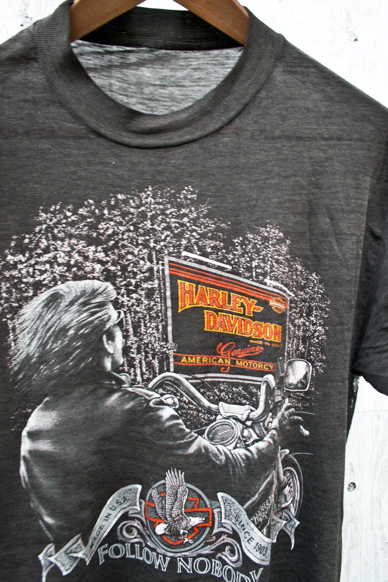 May include: A gray t-shirt with a Harley-Davidson graphic featuring a motorcycle rider in a forest setting. The graphic includes the text "Harley-Davidson", "Genuine American Motorcy", and "Since 1903". The shirt also has the text "Made in USA" and "Follow Nobody" printed on it.