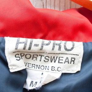 May include: A white tag with black text that reads "HI-PRO SPORTSWEAR VERNON B.C."  The tag is attached to a red and blue jacket.