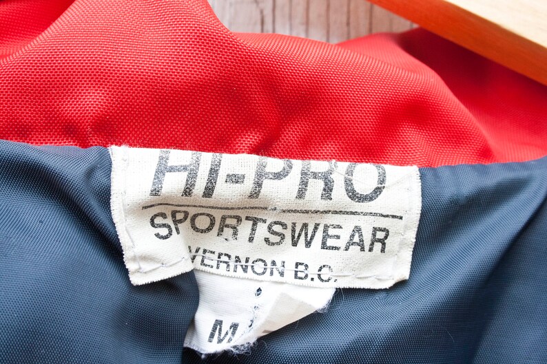 May include: A white tag with black text that reads "HI-PRO SPORTSWEAR VERNON B.C."  The tag is attached to a red and blue jacket.