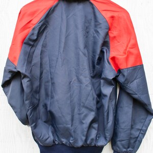 May include: A navy blue and red windbreaker jacket with a zipper closure. The jacket has a stand-up collar and long sleeves.