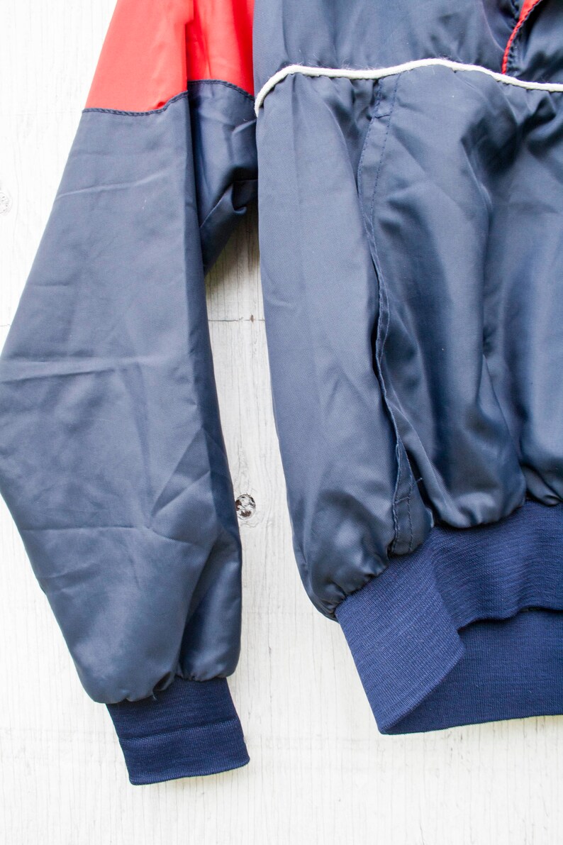 May include: A blue and red windbreaker jacket with a white stripe around the collar and cuffs. The jacket has a zipper closure and a ribbed waistband.