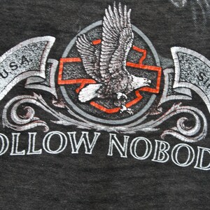 May include: A black t-shirt with a silver eagle graphic and the text "FOLLOW NOBODY" in silver lettering. The eagle is flying over a red cross with a silver circle around it. The text "MADE IN U.S.A." is on a silver banner above the eagle. The text "SINCE 1903" is on a silver banner below the eagle. The text "HARLEY DAVIDSON" is on a silver banner to the right of the eagle.