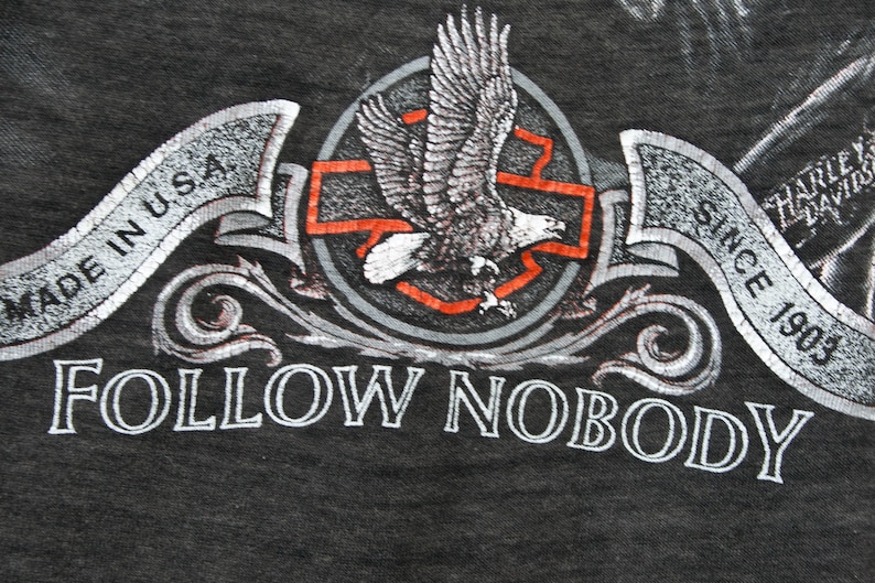 May include: A black t-shirt with a silver eagle graphic and the text "FOLLOW NOBODY" in silver lettering. The eagle is flying over a red cross with a silver circle around it. The text "MADE IN U.S.A." is on a silver banner above the eagle. The text "SINCE 1903" is on a silver banner below the eagle. The text "HARLEY DAVIDSON" is on a silver banner to the right of the eagle.