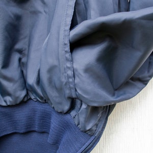 May include: A navy blue jacket with a white zipper and white piping. The jacket has a pocket on the left side and a ribbed waistband.