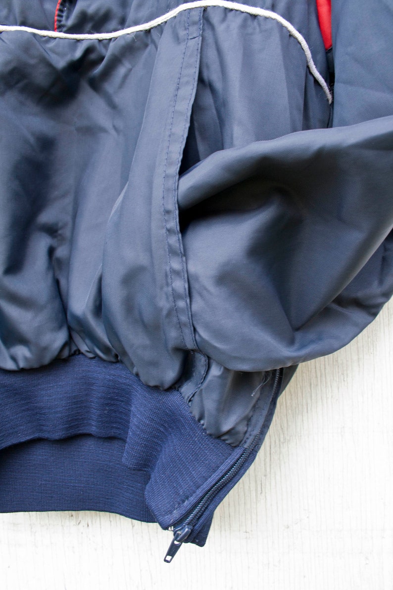 May include: A navy blue jacket with a white zipper and white piping. The jacket has a pocket on the left side and a ribbed waistband.