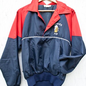 May include: A navy blue and red windbreaker jacket with a white zipper and a white stripe across the chest. The jacket has an embroidered logo that says "Frankies Pub" with a picture of a beer mug.