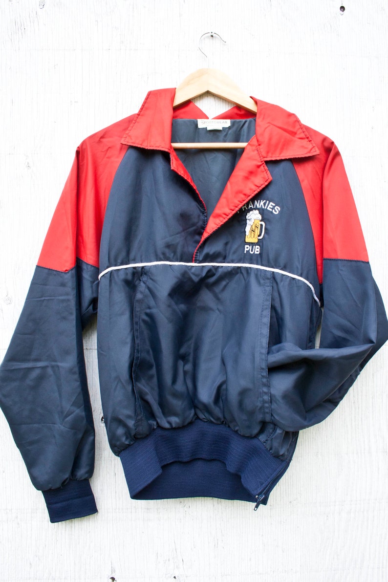 May include: A navy blue and red windbreaker jacket with a white zipper and a white stripe across the chest. The jacket has an embroidered logo that says "Frankies Pub" with a picture of a beer mug.