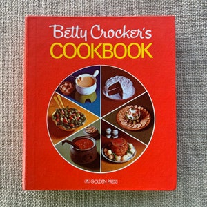 May include: A red cookbook with the title "Betty Crocker's Cookbook" in yellow lettering. The cover features a circular design with various food items, including a cake, a fondue pot, and a salad.