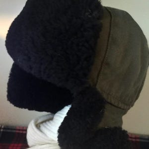 May include: A winter hat with ear flaps. The hat features a dark green fabric crown and black, fluffy ear coverings. A white scarf is visible below the hat. The hat is designed to provide warmth and protection from the cold.