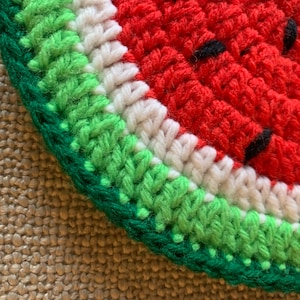 May include: Close-up of a crocheted watermelon with green, white, and red yarn. The watermelon is on a brown burlap background.