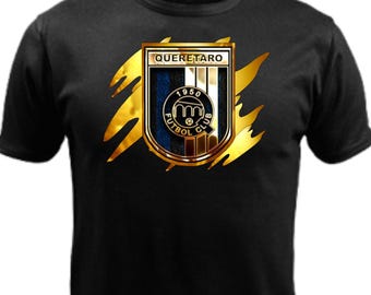 Pode incluir: Uma t-shirt preta de manga curta com um gráfico central do logótipo do Queretaro Futbol Club. O emblema é emoldurado por um design dourado metálico com marcas de garras. O logótipo inclui o texto QUERETARO, 1950 e FUTBOL CLUB.