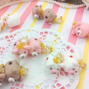 Könnte beinhalten: Eine Sammlung handgefertigter Ohrringe aus Polymer Clay mit niedlichen Tiermotiven. Die Ohrringe sind rosa, braun und weiß mit goldenen Akzenten. Die Ohrringe werden auf einer weißen Spitze mit einem rosa-weiß gestreiften Hintergrund präsentiert.